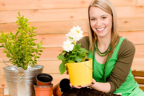 A woman with blonde hair smiling while sitting in a garden area, holding a yellow flower pot with a lush, green flowering plant. In front of her, there are other empty plant pots, including a small black pot, and a large metallic bucket containing a shrub with bright green leaves. The garden features a paved surface, and in the background, a wooden fence with horizontal planks provides a natural, warm backdrop. The scene suggests outdoor gardening activities, with natural daylight illuminating the space, emphasizing the vibrant green foliage and the mixed textures of soil, paving, and plant leaves. This setting reflects a well-maintained outdoor garden area suitable for landscaping and gardening services in Wembley, with attention to plant arrangement and garden aesthetics.